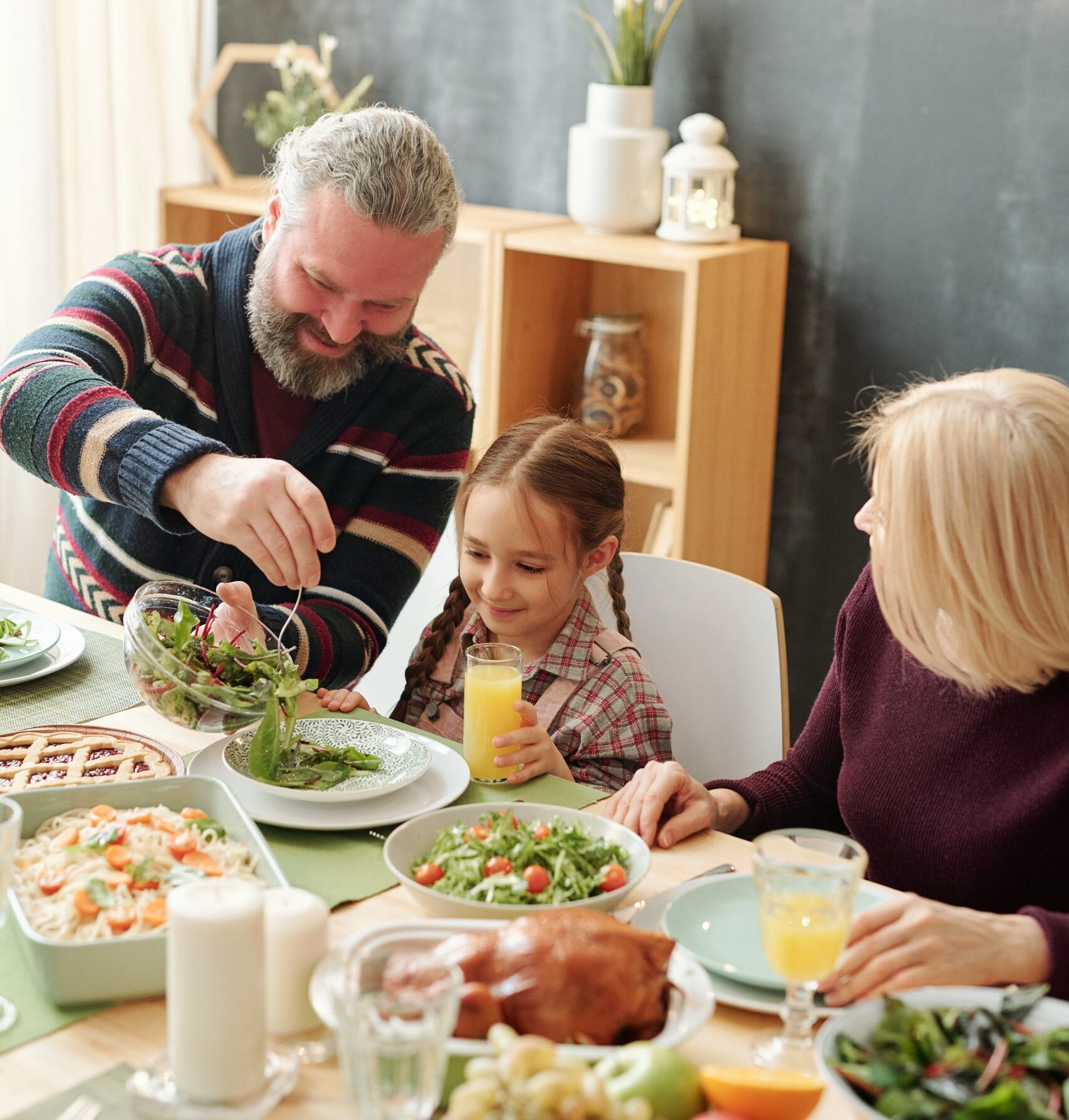 Happy mature man giving salad to his adorable granddaughter by festive table